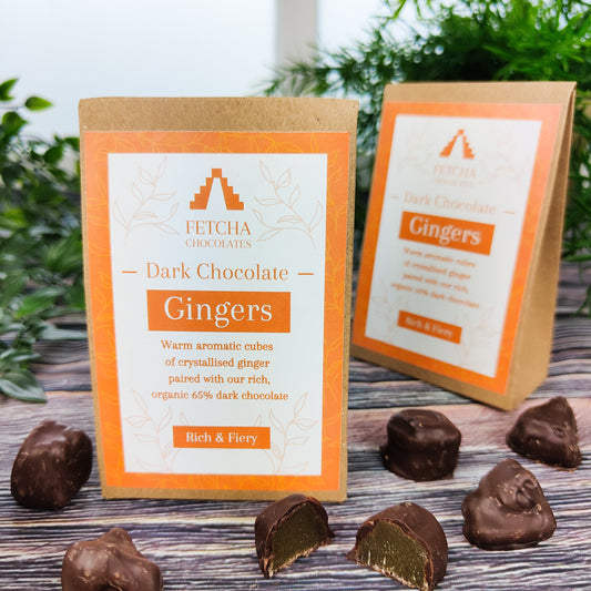 Image shows Fetcha Chocolates handmade dark chocolate gingers,  which are vegan, dairy free, gluten free and come in plastic-free packaging. The packaging is brown kraft cardboard with an orange label. Some dark chocolate gingers are shown outside the box with one cut in half showing the ginger.