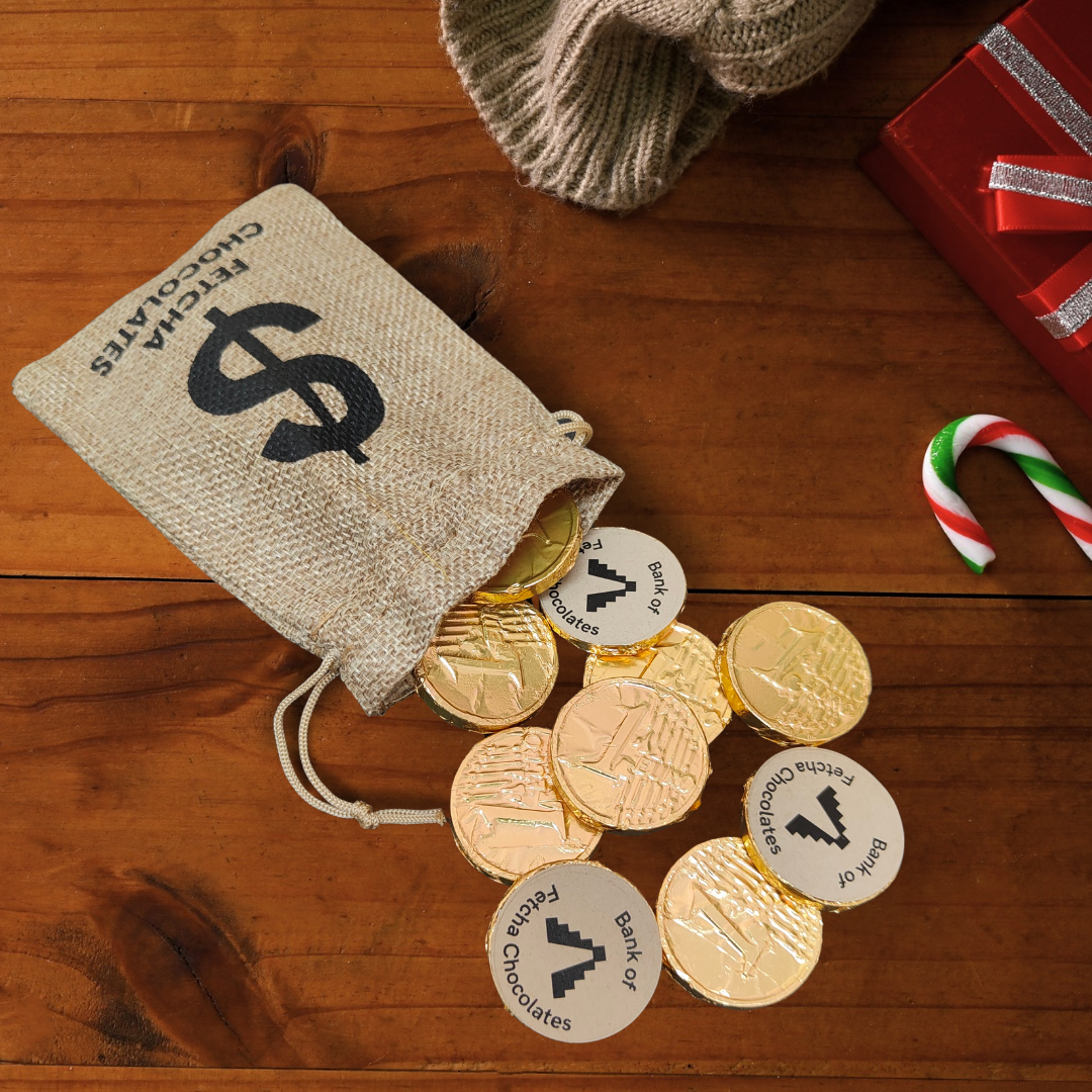 Gold foil wrapped vegan chocolate coins with labels spilling out of a hessian bag on a wooden surface.