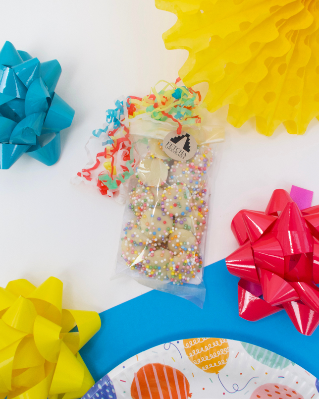 A clear bag containing white chocolate drops with multicoloured sprinkles on a table with decorative ribbons in the background.