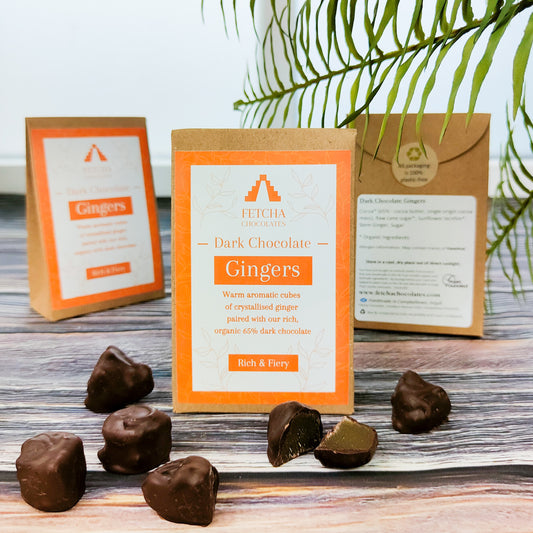 Image shows Fetcha Chocolates handmade dark chocolate gingers,  which are vegan, dairy free, gluten free  and come in plastic-free packaging. The packaging is brown kraft cardboard with an orange label. Some dark chocolate gingers are shown outside the box with one cut in half showing the ginger. 